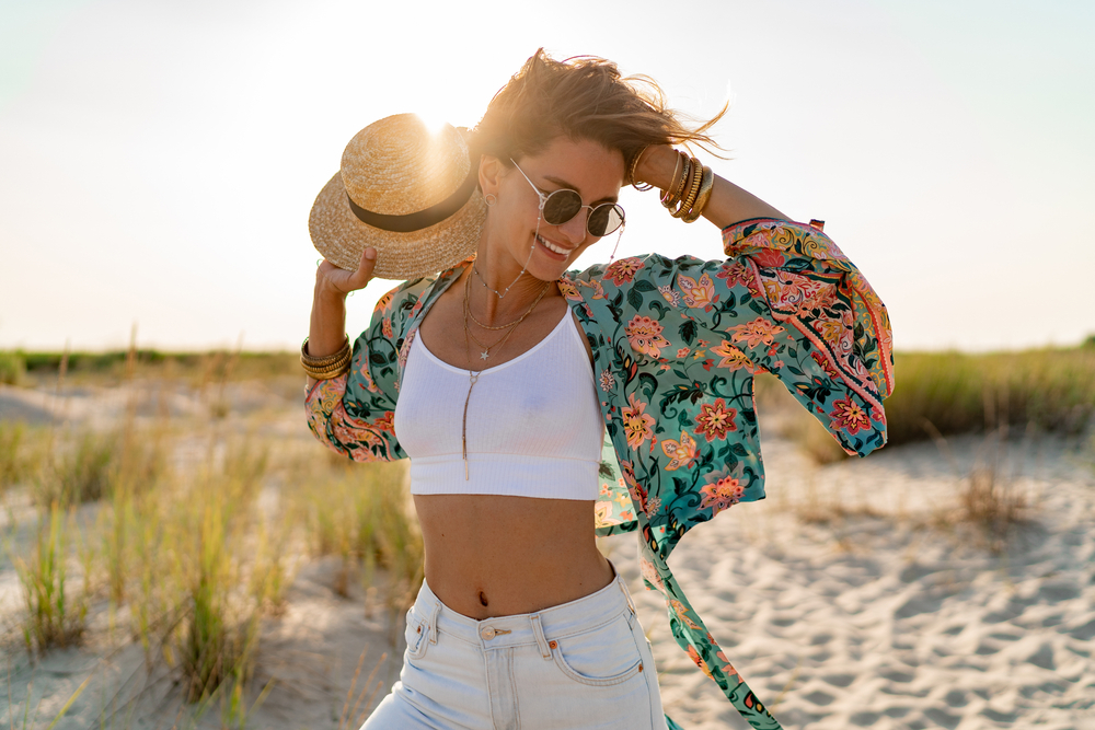 sunny beach in summer style fashion trend outfit happy, freedom, wearing white top, jeans and colorful printed tunic boho style chic and straw hat