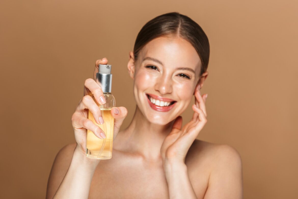 Cheerful and happy young amazing woman with clean and glowing skin posing isolated over brown chocolate background wall holding oil, concept of cleansing oil.