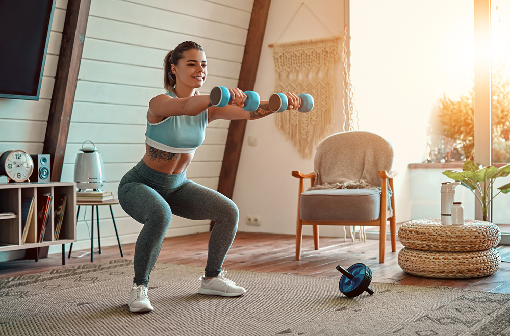 Beautiful young athletic girl in leggings and top crouches with dumbbells at home. Sport, healthy lifestyle.