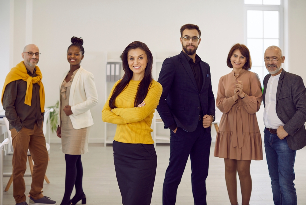 Female business team leader at work. Happy confident young woman in a yellow jumper smiling and looking at the camera standing in the office with a group of employees and teammates in the background