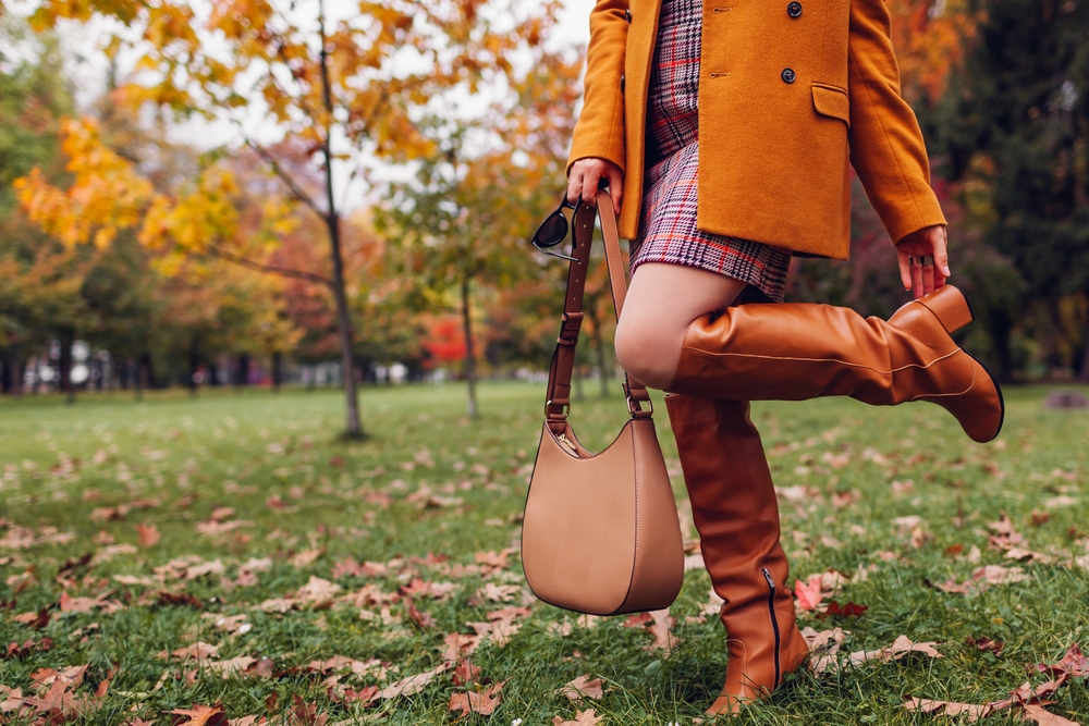 Orange knee high boots. Fashionable woman wearing stylish blazer plaid mini skirt walking in fall park with beige handbag among leaves.