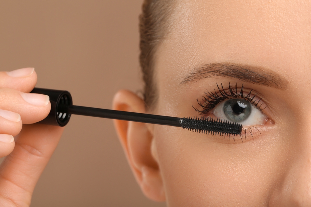 Woman applying mascara onto eyelashes against light brown background, closeup