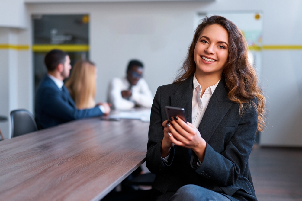 Confident businesswoman in modern office smiles, uses smartphone. Team collaborates in background. Professional woman in suit multitasks, engages in mobile tech communication, team strategy session.