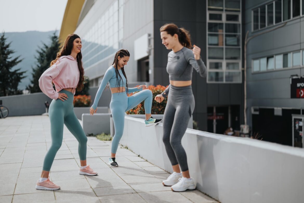 Three young women stretching before a workout, wearing sporty outfits, with an urban backdrop.