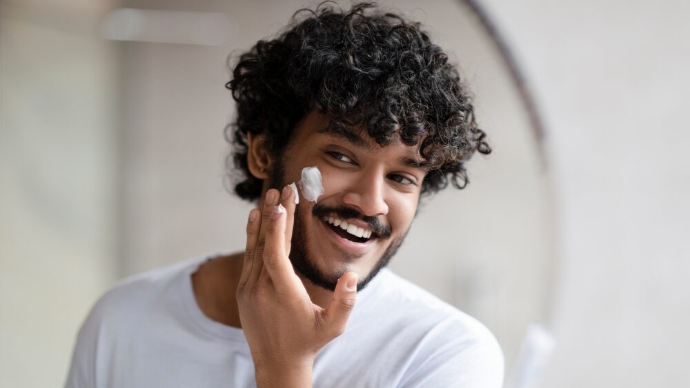 Facial skincare. Indian man applying moisturizing cream on face, looking at his reflection in bathroom mirror at home, free space. Male beauty and skin care