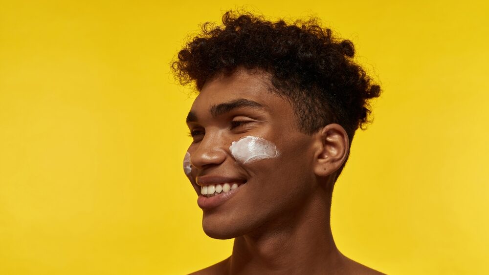 Cropped image of smiling black guy with cosmetic cream on his face looking away. Young brunette curly man. Facial skin care. Isolated on yellow background.