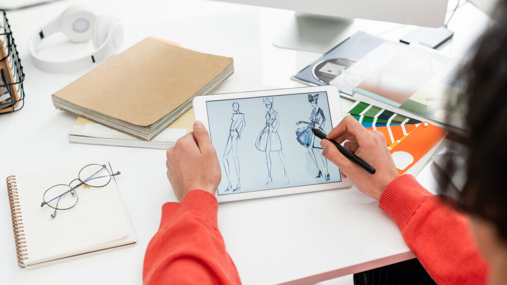 Hands of creative fashion designer with pen pointing at tablet screen while drawing dresses
