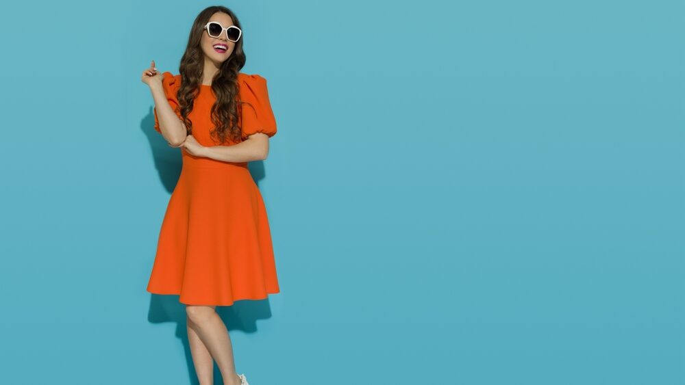 Smiling young woman in orange cocktail mini dress with puffed short sleeves and high heels sandals standing against a blue sunny wall, representing the concept of minimal dressing/fashion
