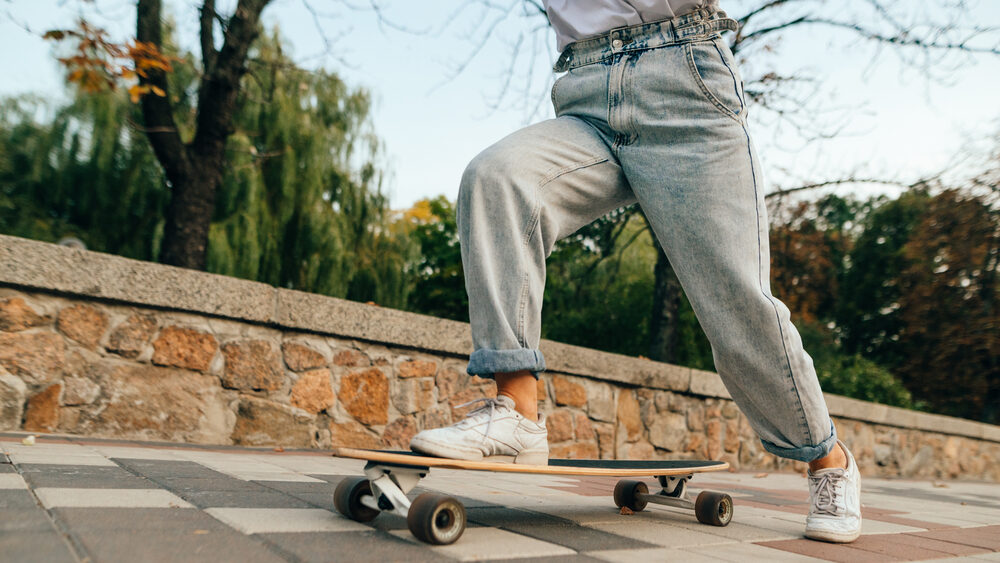 Cropped photo of female skateboarder legs in baggy jeans, riding her long board on a city street near the park.