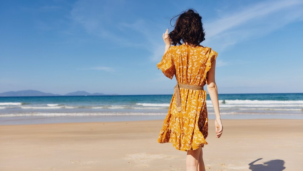 Woman standing on the beach in a yellow A-line dress from 1950 fashion looking out at the vast ocean and sky, contemplating nature's beauty